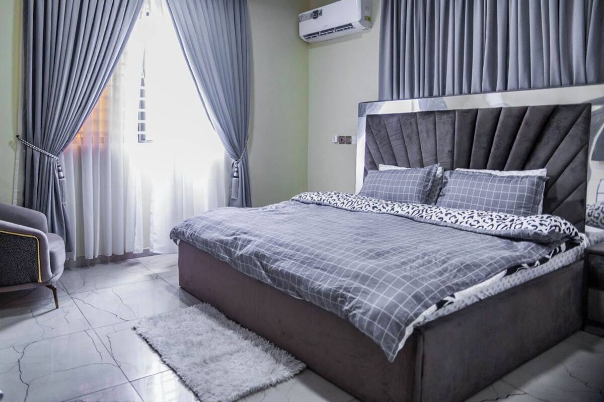 A modern bedroom with a large bed, grey curtains, an air conditioning unit, and a grey sofa.