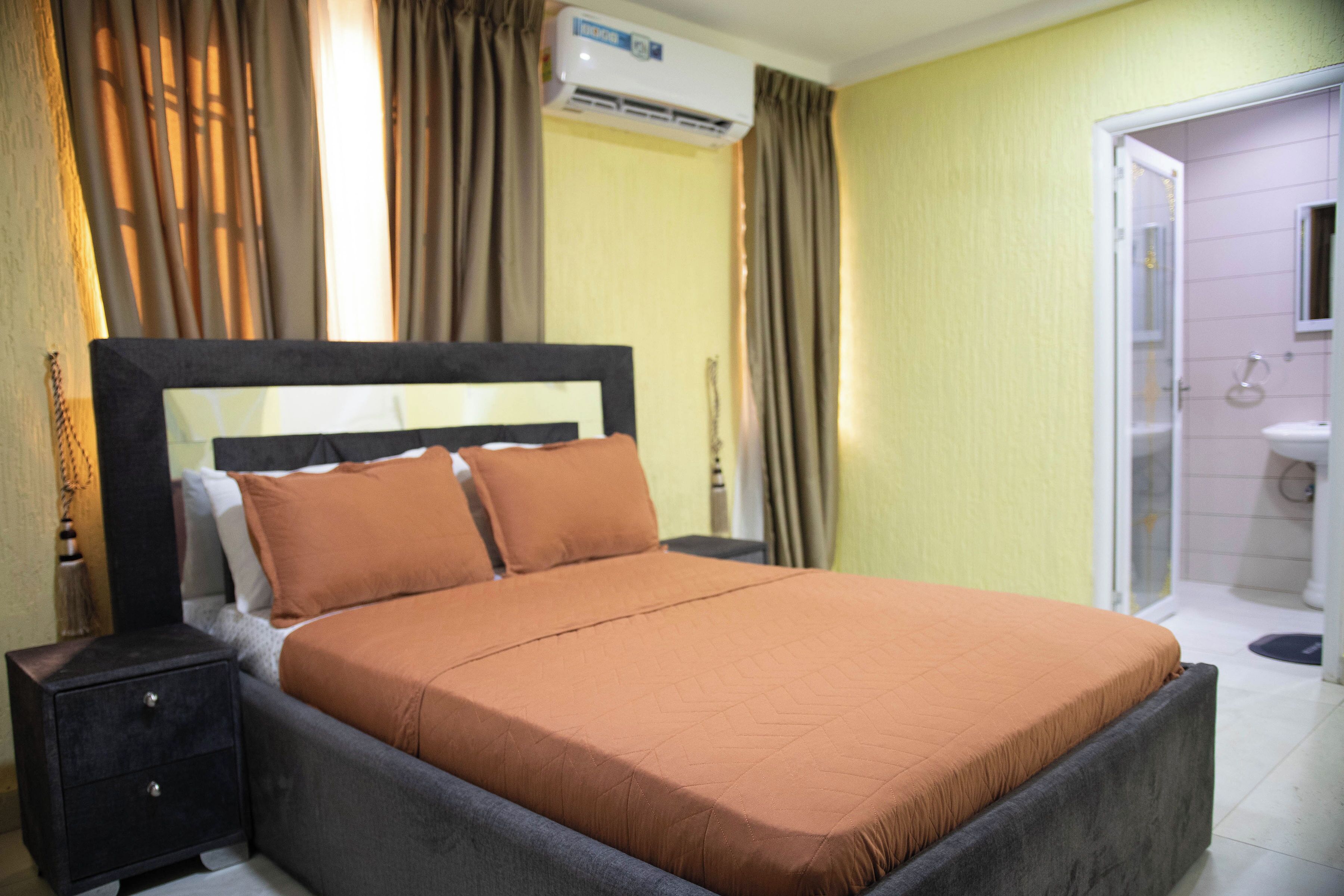 Habitación de hotel con una cama, una mesita de noche, equipo de aire acondicionado y un baño visible a través de una puerta abierta.