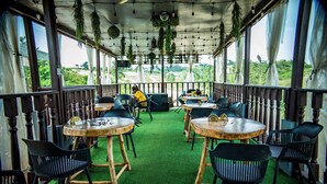 An outdoor dining area with round tables and chairs, surrounded by a wooden railing, and overlooking a scenic landscape.