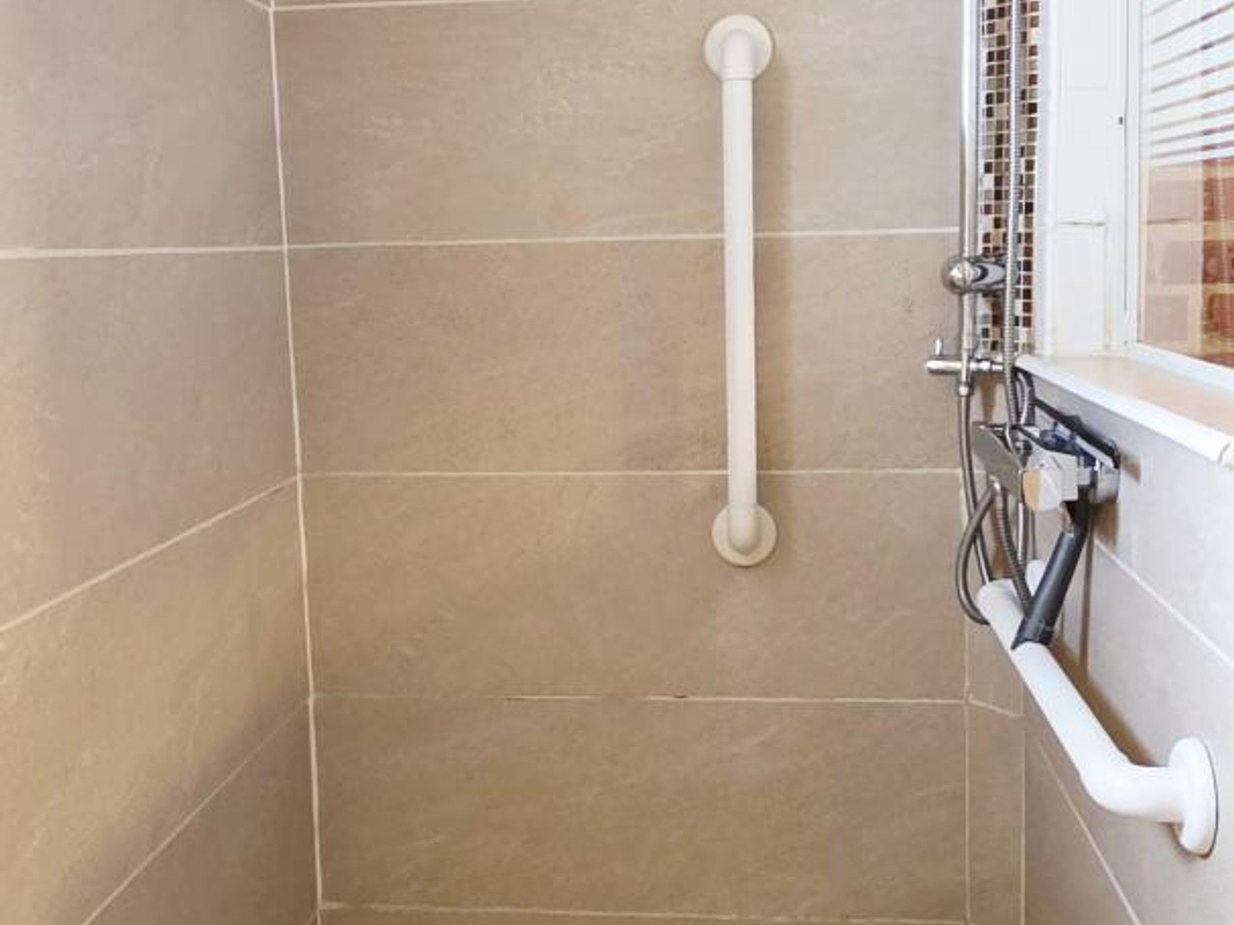 Une salle de bain avec des carreaux de couleur beige, une barre d’appui blanche et un panneau de commande de douche.