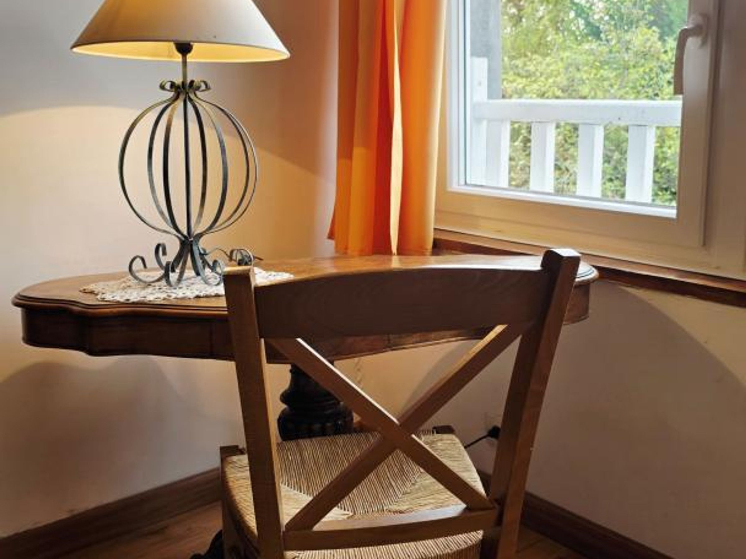Une chaise en bois, dotée d’un siège et d’un dossier en osier, est placée devant une table sur laquelle se trouve une lampe. La table est située près d’une fenêtre ornée de rideaux.