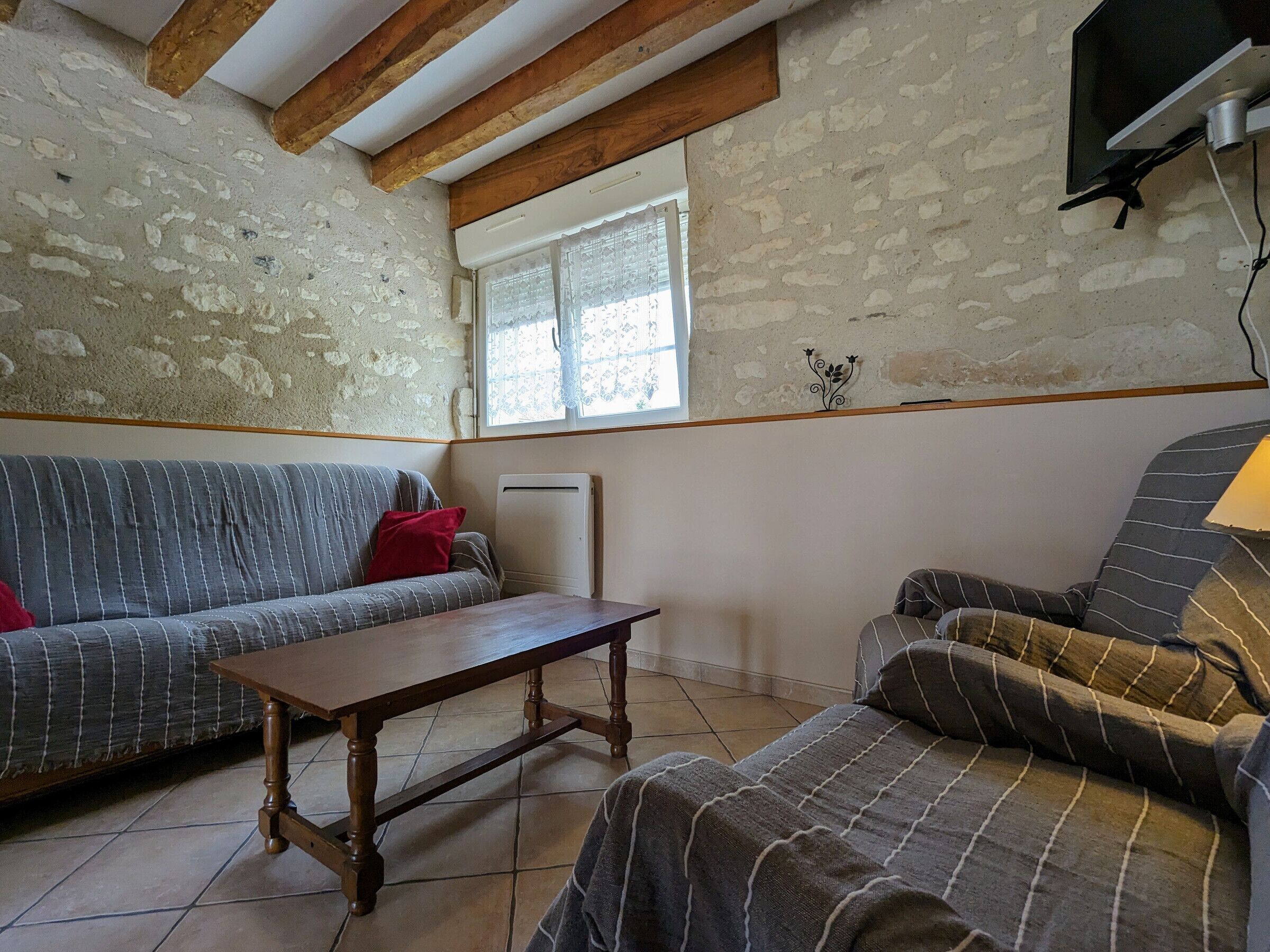 A room with a stone wall, a wooden ceiling, a window, a sofa, a chair, and a TV mounted on the wall.