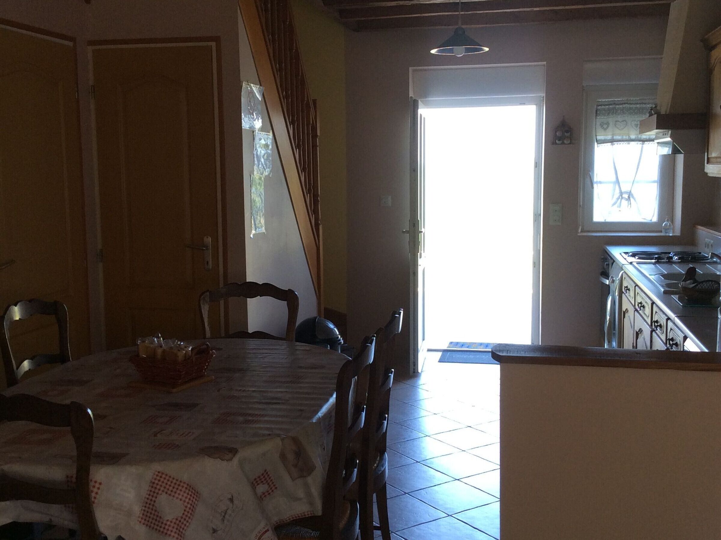 Un coin repas avec une table dressée pour quatre personnes, un escalier et une cuisine visible à travers une porte ouverte.