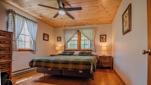 A bedroom with a bed, wooden floor, ceiling fan, two windows with curtains, bedside lamps, and a door. - Glatts - Cabin located in Shunk (Shunk)