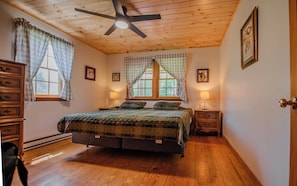 Un dormitorio con cama, suelo de madera, ventilador de techo, dos ventanas con cortinas, lámparas de noche y una puerta.