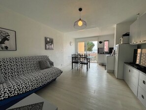 Ein modernes Wohnzimmer mit einem gemusterten Sofa, einem Essbereich mit Stühlen sowie einer Küche mit Schränken und Geräten.