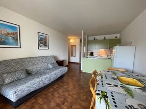 A living room with a sofa, a dining table with a chair, a kitchen with a refrigerator, and two framed pictures on the wall. - Apartment with parking - 6 guests (Cap d'Agde)