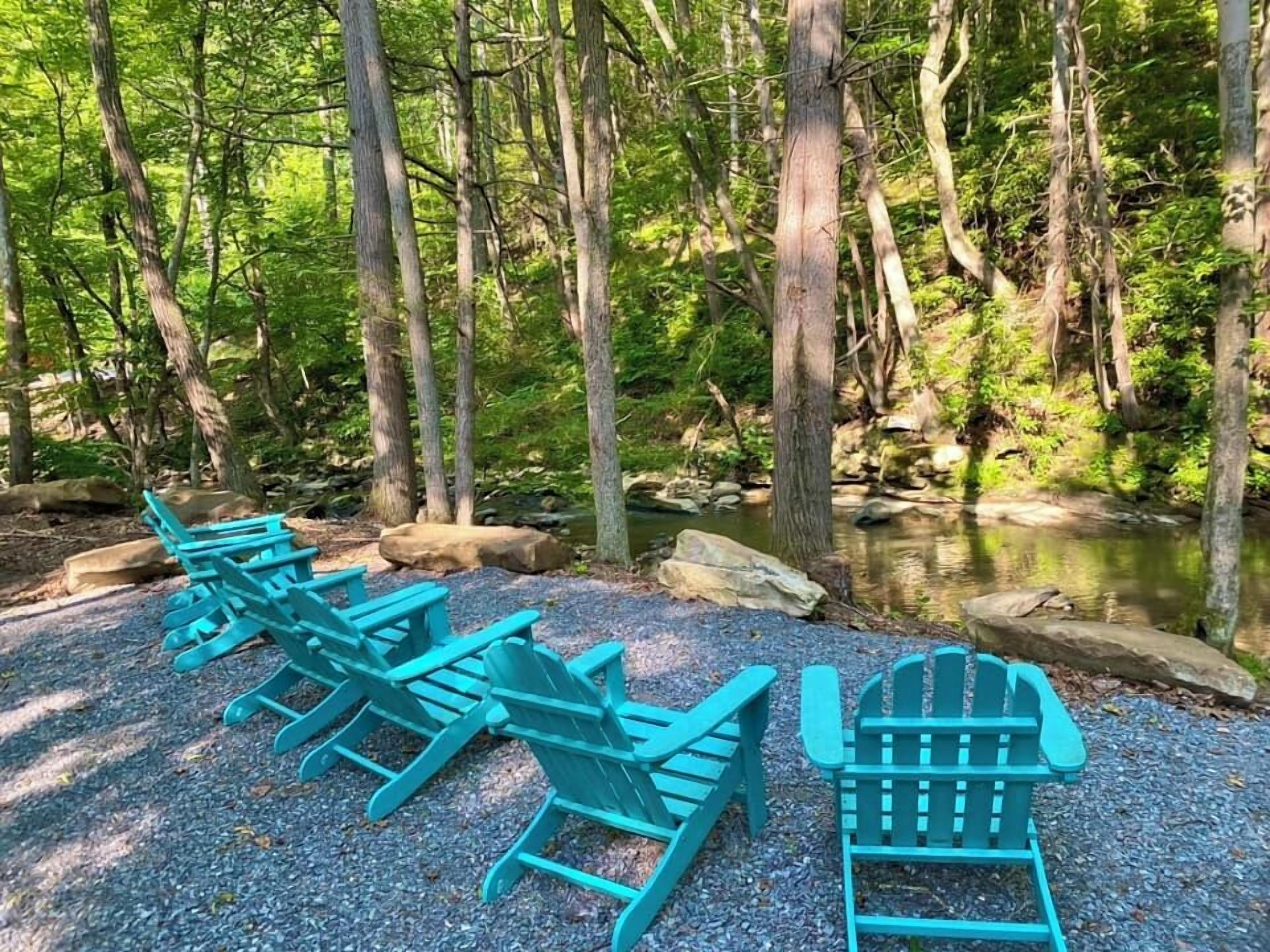 A row of turquoise Adirondack chairs facing a forested area with a stream.