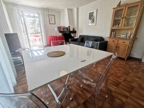 Ein Essbereich mit einem weißen Tisch und Stühlen, ein roter Tisch im Hintergrund, eine schwarze Couch und eine Holzkabine mit Glastüren.