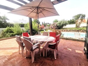 Eine Terrasse mit Tisch, Stühlen und Sonnenschirm, mit Blick auf ein Schwimmbad und Grünflächen.