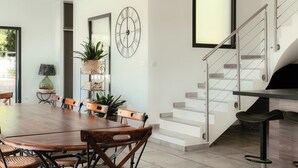 Un área de comedor moderna con una mesa y sillas de madera, una escalera con barandillas de vidrio y un reloj de pared grande.