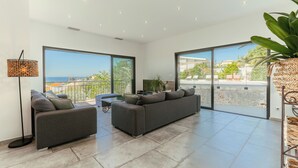Amplio salón con un sofá gris grande, una mesita y una lámpara de pie. Grandes ventanales ofrecen vistas al mar y un balcón con mobiliario exterior.