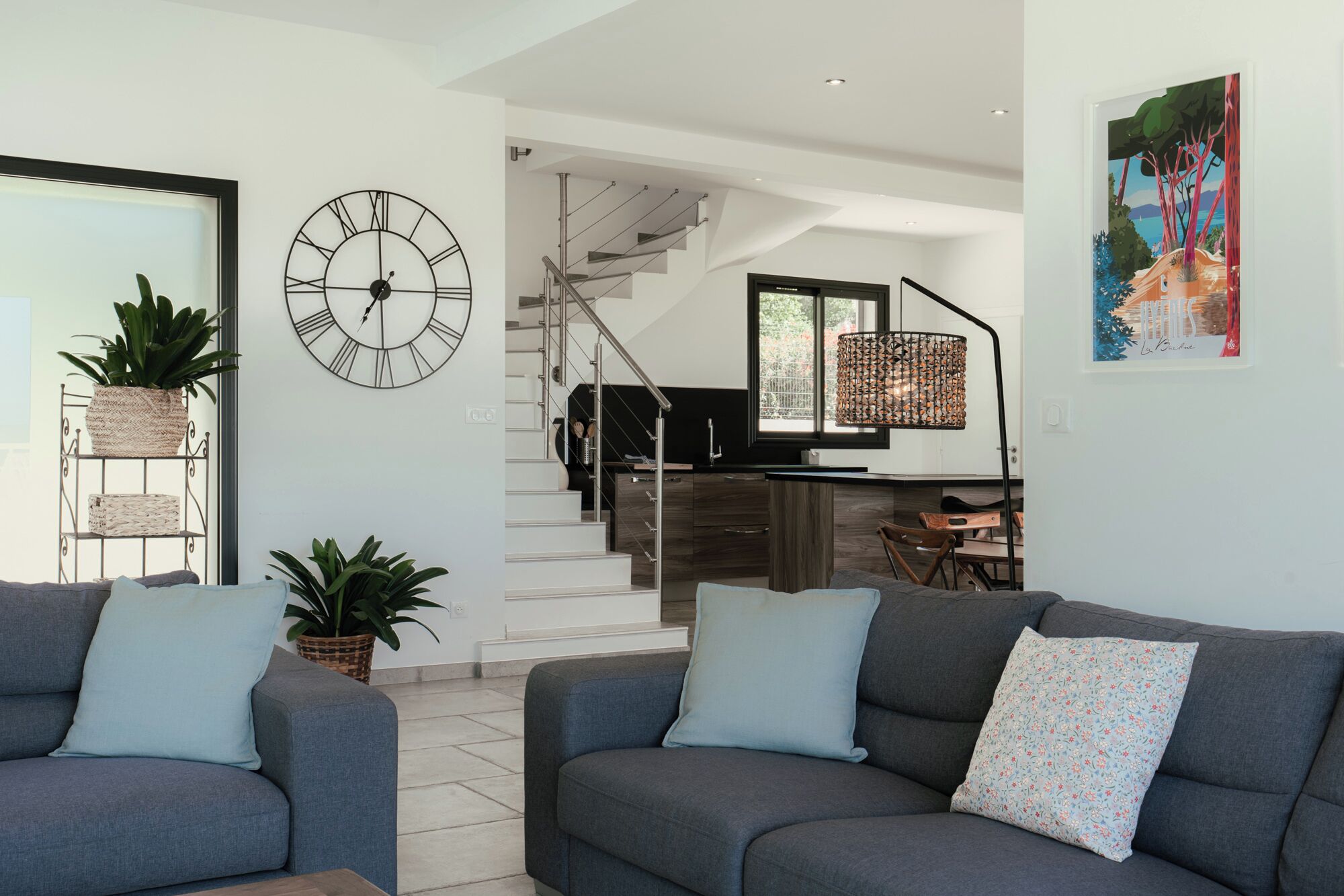 Ein modernes Wohnzimmer mit einem grauen Sofa, einer großen Wanduhr und einer Treppe, die in einen Küchenbereich führt.