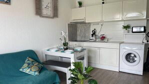 A compact living space with a kitchenette, a washing machine, a dining table with chairs, a sofa, and a clock on the wall. - Studio with AC and pool - 4 guests (Cap d'Agde)