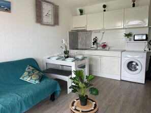 Ein kompakter Wohnraum mit Küchenzeile, einer Waschmaschine, einem Esstisch mit Stühlen, einer Couch und einer Uhr an der Wand.