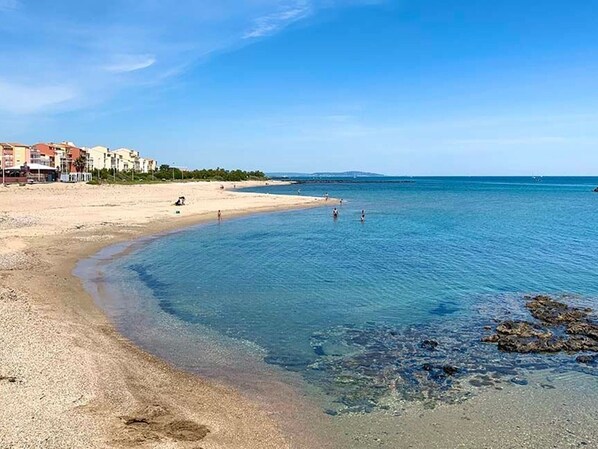 Ein Strand mit klarem Wasser, sandigem Ufer und Gebäuden im Hintergrund.