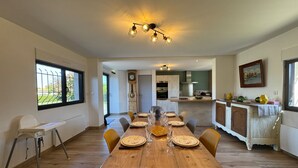 Un coin repas avec une table en bois dressée pour six personnes, une cuisine en arrière-plan et une grande fenêtre donnant sur un paysage verdoyant.