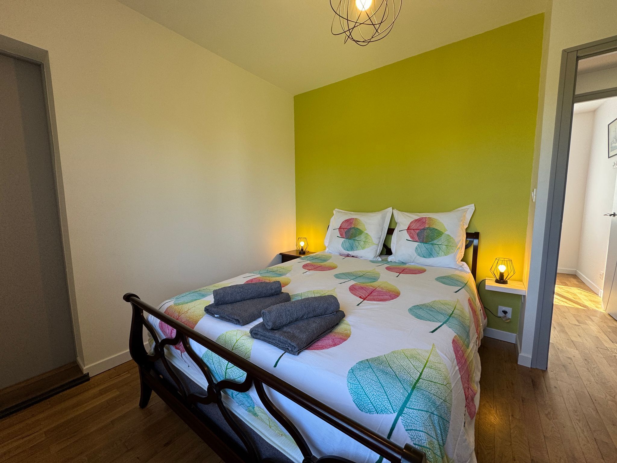 Un lit bien fait, recouvert d’une couvre-lit à motifs de feuilles, avec deux lampes de chevet et une tête de lit en bois.