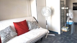 En hvid sofa med mønstrede puder, en gulvventilator, en metalkurv med ventilator og et flisegulv.