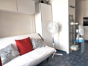Ein weißes Sofa mit gemusterten Kissen, ein Standventilator, ein Metallregal mit Ventilator und ein Fliesenboden.