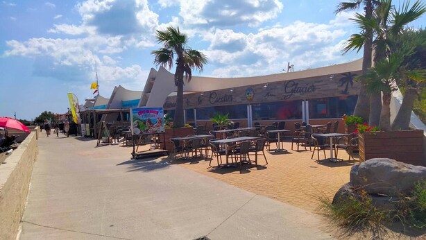 Un restaurante frente a la playa con terraza, palmeras y cielo despejado.