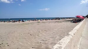 Una playa con un muro de hormigón, gente y barcos a lo lejos.