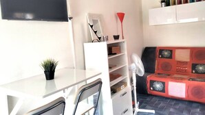 Un espacio habitable compacto con un escritorio blanco, una silla, una planta pequeña, un sofá estampado en rojo y negro, un ventilador blanco y una estantería con varios objetos.