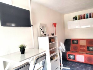 A compact living space with a white desk, a chair, a small plant, a red and black patterned sofa, a white fan, and a shelf with various items. - Ideal studio in Roquille - 4 guests (Cap d'Agde)