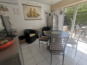 A dining area with a round table and chairs, a black sofa, a refrigerator, and a window with a view of greenery. - Apartment with pool access - 4 guests (Cap d'Agde)