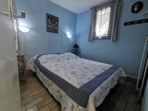 A bedroom with a bed, a nightstand, a lamp, a window with curtains, and a wall-mounted picture. - Quiet terraced house - 4 guests (Cap d'Agde)