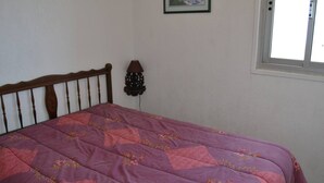 Un dormitorio con cabecera de cama de madera, colcha de flores moradas, lámpara de pared e imagen enmarcada sobre la cama.