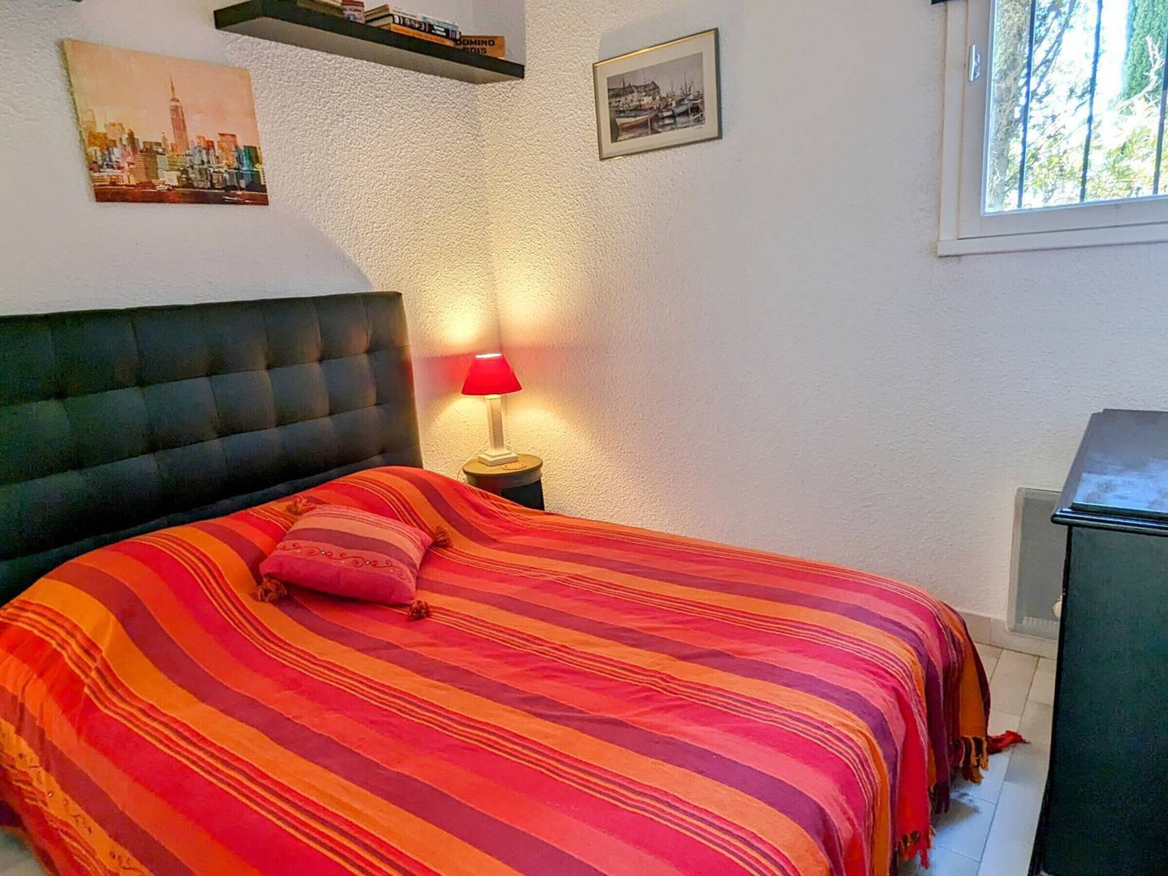 Une chambre à coucher avec un lit, une table de chevet avec une lampe, un tableau représentant un paysage urbain, un tableau encadré et une fenêtre.