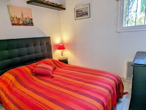A bedroom with a bed, a nightstand with a lamp, a cityscape painting, a framed picture, and a window. - Apartment with air conditioning - 4 guests (Cap d'Agde)