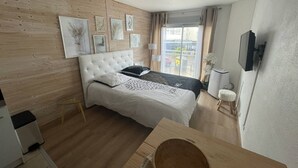 A bedroom with a bed, a wooden headboard, a window with curtains, a small table, and a wall-mounted TV. - Studio with port view - 2 guests (Cap d'Agde)