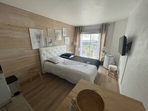 A bedroom with a bed, a wooden headboard, a window with curtains, a small table, and a wall-mounted TV. - Studio with port view - 2 guests (Cap d'Agde)