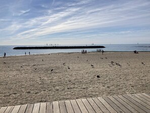 Ein Strand mit einem hölzernen Promenadenweg, sandigem Ufer und Segelbooten in der Ferne.