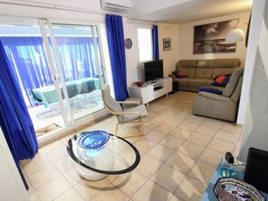 Ein Wohnzimmer mit Glastisch, einer Couch, einem Fernseher und einem Balkon mit blauen Vorhängen.