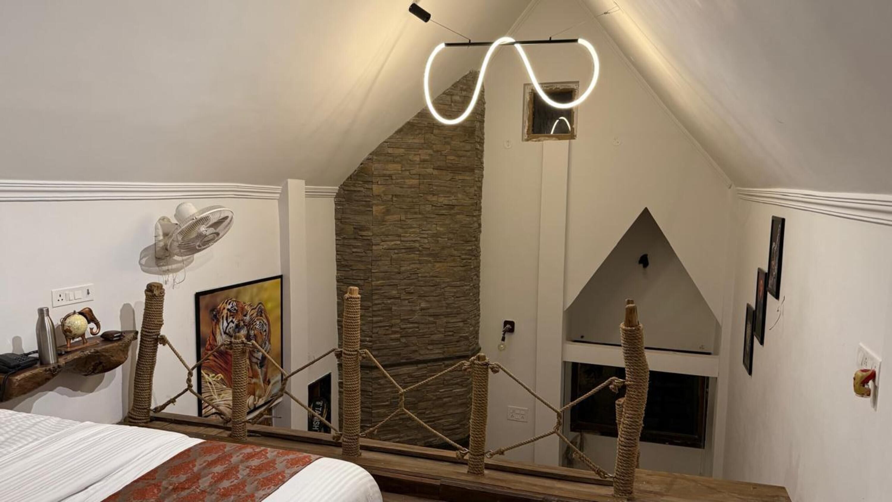 A bedroom with a sloped ceiling, a stone wall, a bed with a patterned bedspread, a fan, a globe, and a framed tiger picture.