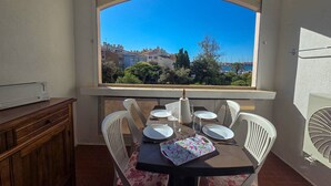 A dining area with a table set for four, a window with a view of buildings and trees, and a white fan on the right. - Air-conditioned apartment at the port - 4 guests (Cap d'Agde)