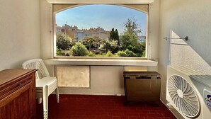 A room with a window overlooking a residential area, a small table, a chair, a radiator, and a trash bin. - Air-conditioned apartment at the port - 4 guests (Cap d'Agde)