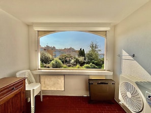 Ein Zimmer mit Fensterblick auf eine Wohngegend, einem kleinen Tisch, einem Stuhl, einem Heizkörper und einer Mülltonne.