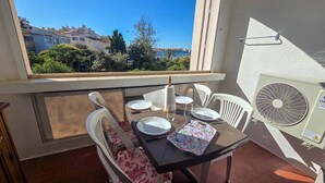 A balcony with a table set for two, white chairs, a fan, and a view of buildings and trees. - Air-conditioned apartment at the port - 4 guests (Cap d'Agde)