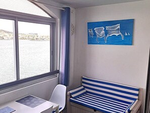 A room with a window, a bench, a table, and a wall painting. - Apartment with port view - 4 guests (Cap d'Agde)