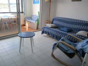 A room with a blue sofa, a small round table, a floor lamp, and a view through a glass door. - Apartment with port view - 4 guests (Cap d'Agde)