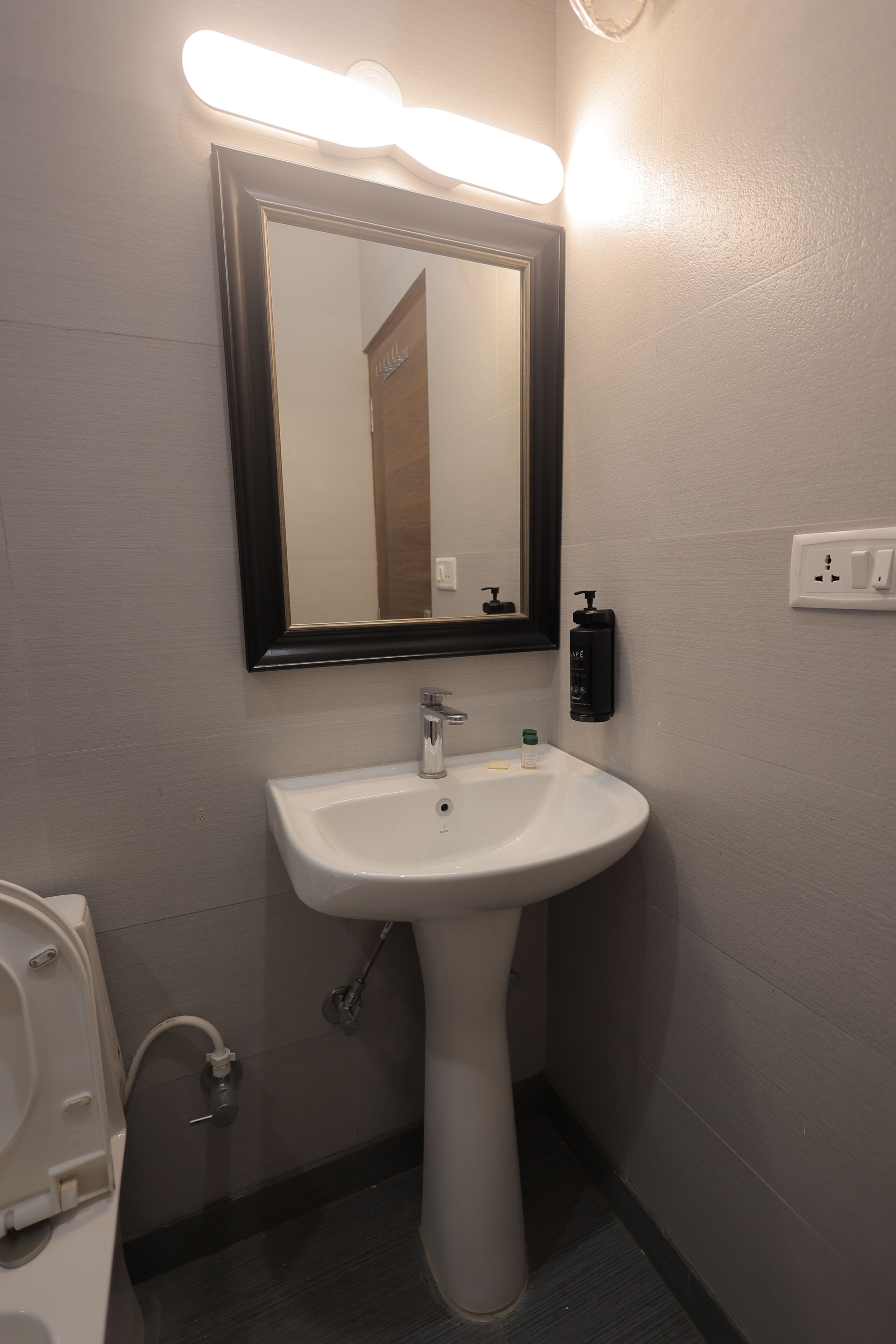 Une salle de bain avec un lavabo blanc, un miroir avec éclairage intégré, un distributeur de savon et des toilettes.
