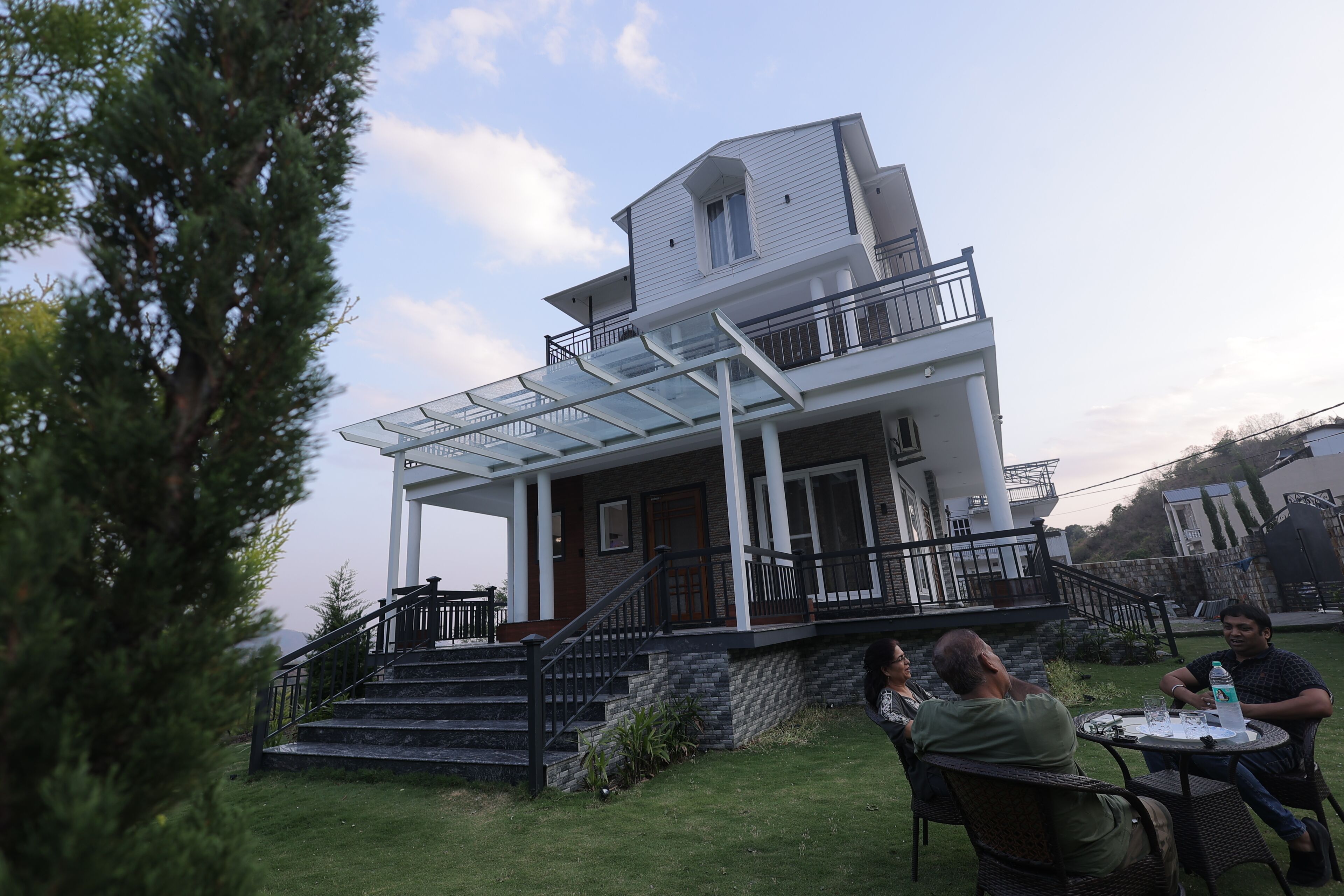A two-story house with a balcony, a staircase, and a patio area where people are seated.
