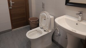 A bathroom with a toilet, bidet, sink, and a wooden door. - Maktain Glance (Dehradun)