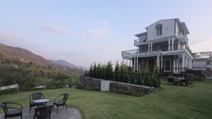 A two-story building with a balcony, a patio with chairs, and a view of a hilly landscape. - Maktain Glance (Dehradun)