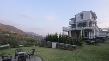 Un edificio de dos pisos con un balcón, un patio con sillas y vistas a un paisaje montañoso.
