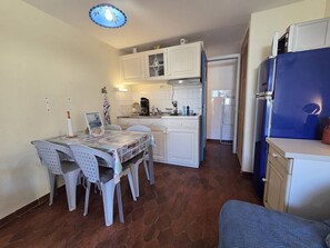 A compact kitchen with white cabinets, a dining table with chairs, a blue refrigerator, and a tiled floor. - Studio with AC, beach access - 3 guests (Cap d'Agde)